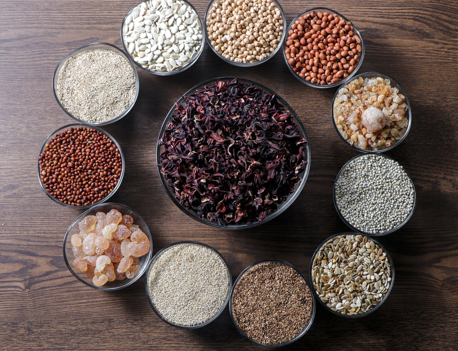 Bowls of our products arranged in a circle on a wooden table: gum arabic, sesame, groundnut, pulses, grains and seeds around a large bowl of dried hibiscus