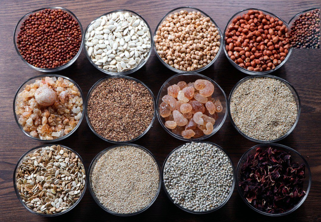 Bowls of our products in rows on a wooden table: gum arabic, sesame, groundnut, hibiscus, watermelon seeds, pulses and grains